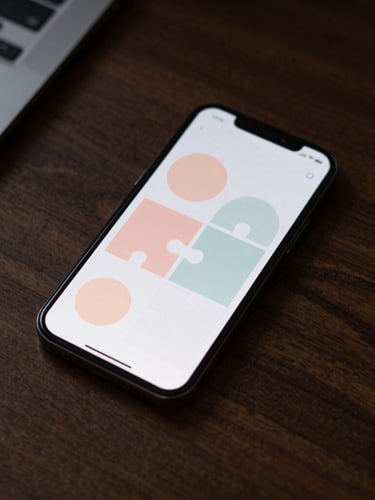 Overhead flat-lay of a dark wooden desk, a modern smartphone displaying a minimalist puzzle game with soft pastel shapes, warm evening light.
