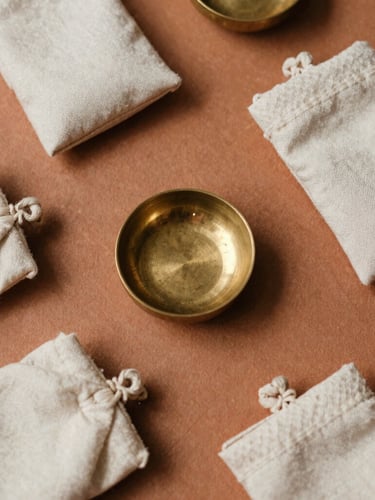 Overhead flat-lay of hand-woven raw silk pouches and small, heavy-milled brass incense holders on a warm clay surface, soft diffused lighting.