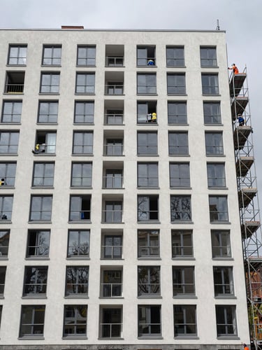 Wide environmental shot of a multi-storey residential building facade, newly installed aluminum window frames visible across multiple floors, overcast daylight, construction crew on scaffolding at right frame, building exterior in a Central European urban setting, documentary framing