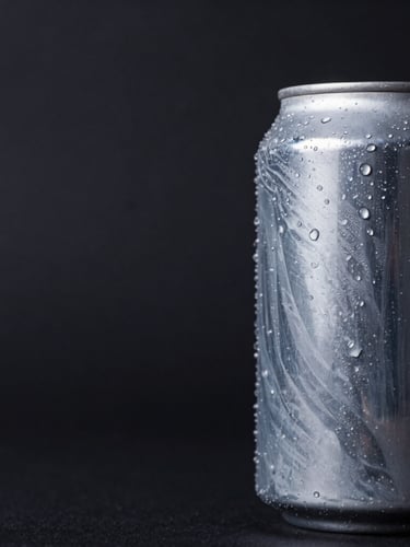 Wide panoramic 21:9 frame, macro close-up of a printed aluminum can positioned to the right third, showing tactile ink textures and condensation beads, dramatic high-contrast studio lighting against a deep charcoal background.