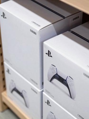 Overhead close-up study of stacked sealed Sony PlayStation 5 retail boxes on a warehouse shelf, authentication seal labels visible on box edges, clean studio strobe lighting revealing barcode and serial number stickers, matte white packaging, no hands or people in frame