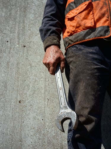 Low-angle heroic close-up of a worker in high-vis gear holding a heavy iron wrench, weathered hands, harsh daylight, concrete background.