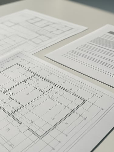 A close-up of physical blueprints and structured compliance documentation on a clean desk under bright daylight.