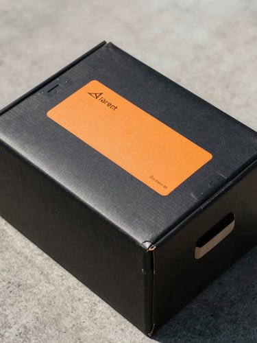A top-down view of a sealed, matte-black delivery box with a clean orange transit label on a concrete surface, bright daylight, sharp shadows.