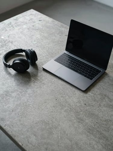 Wide environmental shot of a minimal workspace — matte black laptop open beside a pair of over-ear headphones on a concrete surface, cool north-facing daylight, overhead technical perspective, no people