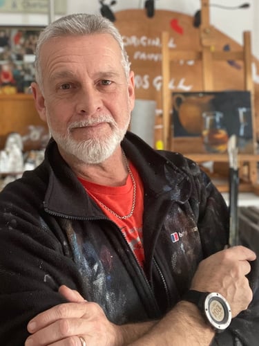 A male professional artist with a grey beard in his art studio with paint-stained clothes and an easel.