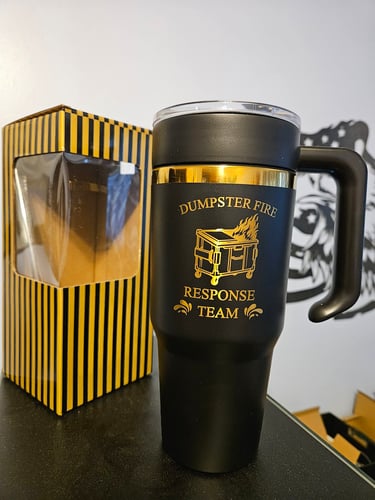 Black and gold Dumpster Fire Response Team insulated travel mug with handle next to a yellow striped gift box.