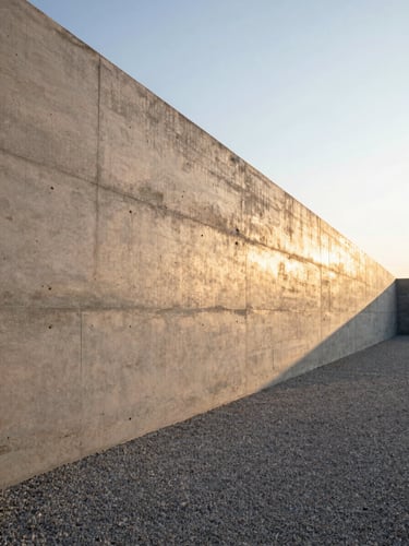 Low-angle golden hour sunlight highlighting the linear texture of a board-formed concrete retaining wall, clean architectural lines framing a minimalist gravel path, sharp shadows.