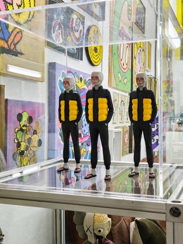 Three designer vinyl figures in a glass display case with colorful pop art paintings in the background.