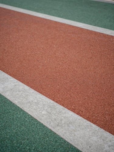 Low-angle architectural shot of a modern playground floor, textured terracotta and forest green rubber tiles meeting clean concrete edges under soft overcast daylight, macro texture visible.