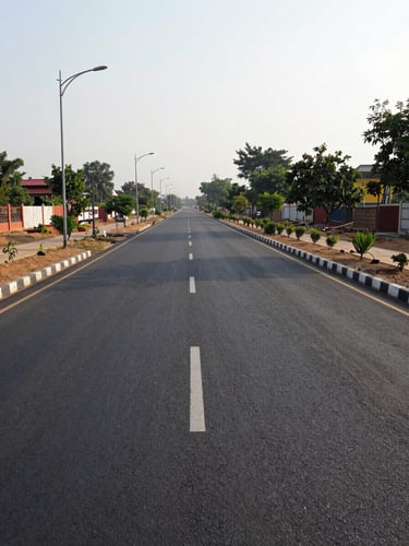Ground-level wide shot along a newly paved internal township road in Jhajjar, smooth asphalt extending into the distance with planted median strip to the right, functional streetlights lining both sides, open sky above, natural afternoon daylight, no people, documentary framing