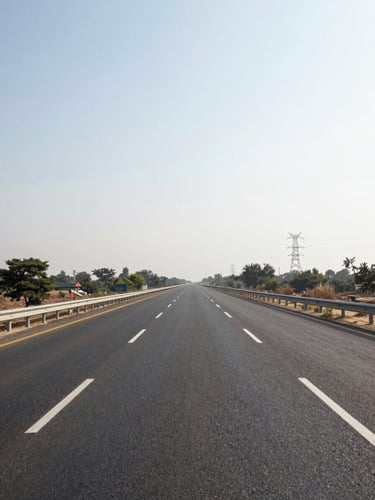 Ground-level view along a wide four-lane highway in Haryana at midday, road markings sharp and clear, flat terrain flanking both sides, power transmission towers visible in the far right distance, overcast bright sky, no vehicles in foreground