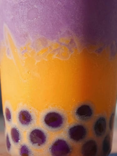 Close-up of a vibrant purple and orange layered bubble tea in a clear cup, textured tapioca pearls visible at the bottom, bright natural daylight with neon accents, 35mm lens