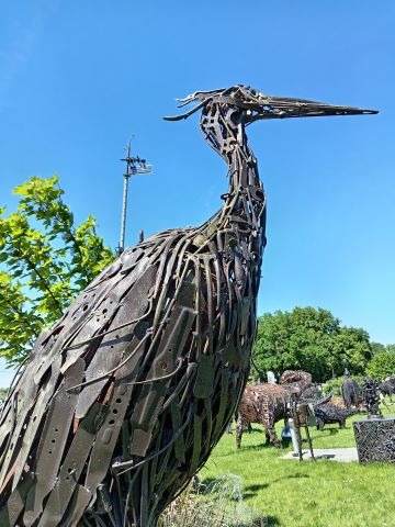 A large outdoor scrap metal heron sculpture standing in a green field under a clear blue sky.