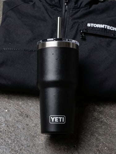 Close-up overhead shot of a matte black Yeti tumbler and a folded Stormtech jacket laid flat on a dark concrete surface, even studio lighting with sharp shadows, no warmth, product labels visible at the edges