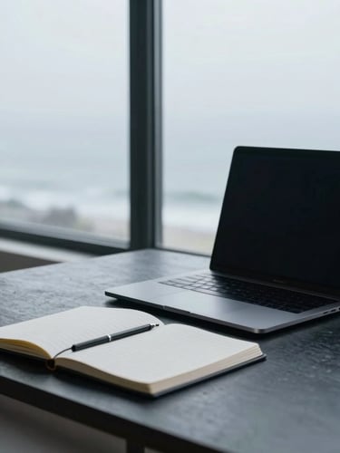 A minimalist workspace in cool, diffused daylight. A sleek modern laptop sits next to a neatly stacked physical notebook on a dark slate desk, with soft coastal fog visible through a large window in the background.