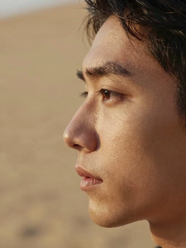 Nahaufnahme eines Gesichts im Profil, weiches natürliches Seitenlicht, ruhiger und neutraler Ausdruck, warmer Sandhintergrund.