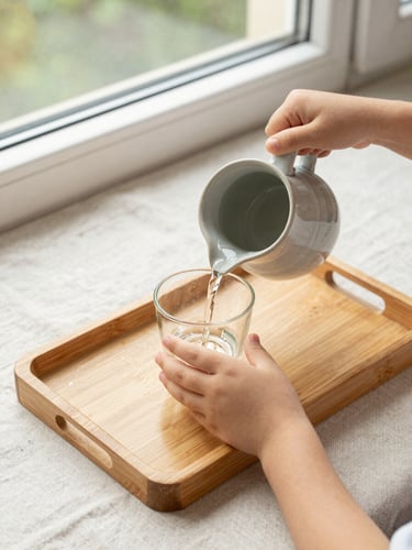 Close overhead view of a child's small hands carefully pouring water from a ceramic pitcher into a glass on a wooden Montessori tray, natural north-facing daylight from a window, materials arranged neatly on a light linen surface, no adult hands visible