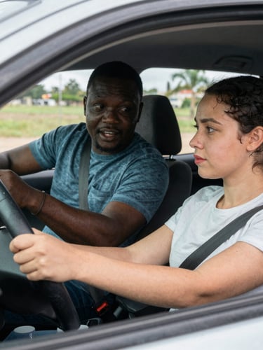 A medium close-up of a patient driving instructor calmly guiding a focused teenage driver inside a stationary training vehicle, soft natural Maryland daylight filtering through the window, warm expressions.