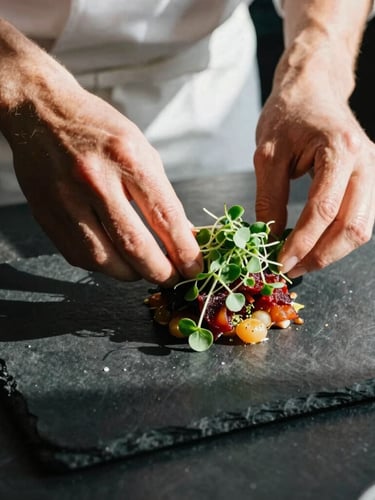 Overhead close-up of a chef's hands carefully plating a vibrant dish with microgreens, dramatic side lighting casting long shadows on a dark slate counter, 35mm