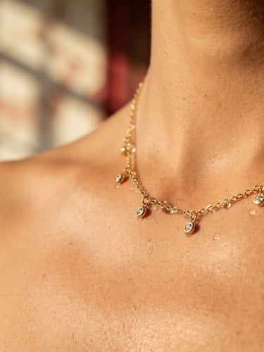 Macro close-up of a delicate gold-plated stainless steel necklace resting on warm skin, water droplets glistening on the metal, soft golden hour sunlight, architectural shadows