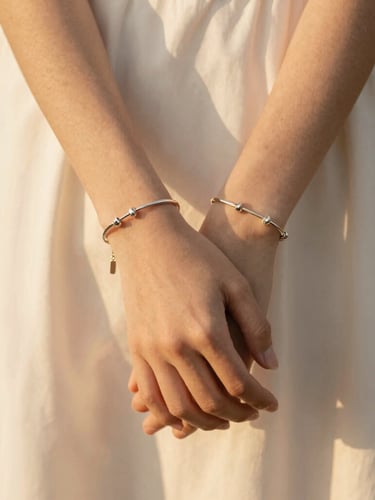 Close-up portrait of hands clasped, showing delicate steel bracelets catching warm golden hour sunlight, soft cream fabric background, minimalist editorial composition.
