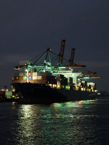 Cinematic wide-angle view of a massive container ship docked at a high-tech automated port at twilight, glowing green terminal lights, deep obsidian water, dramatic backlighting.