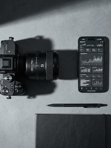 Minimalist modern workspace, high-contrast studio lighting, sharp shadows, overhead desk layout with a professional camera lens and a smartphone displaying clean analytics charts, monochrome tones.