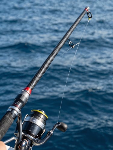 Extreme close-up of a heavy carbon fishing rod bent under extreme load, high-contrast midday sun reflecting off deep blue ocean water, water spraying from the line.