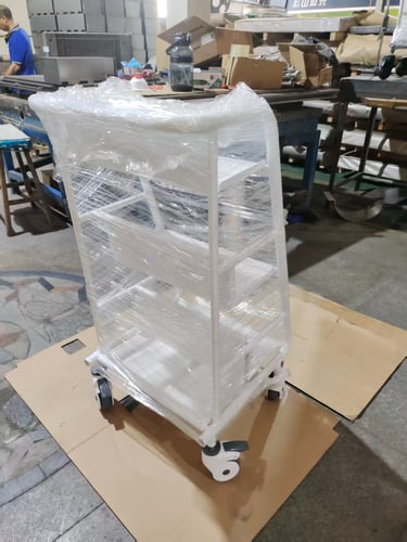 White metal rolling storage cart with shelves wrapped in plastic film for shipping in a factory.