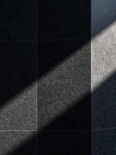 Abstract architectural crop of a monolithic black granite structure, sharp sunlight cutting across raw stone, deep shadows, high-contrast minimalist composition.