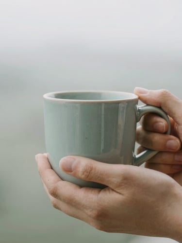 A close-up of hands holding a warm ceramic mug in diffused overcast daylight, soft muted sage background, shallow depth of field, 35mm photography.