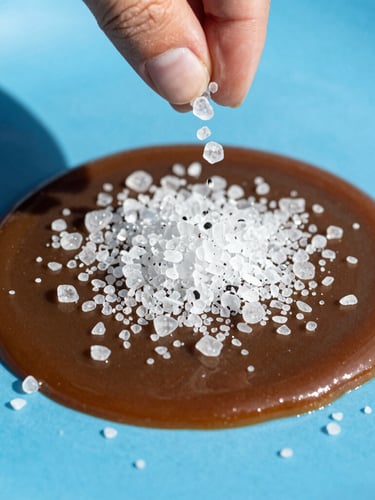 Close-up of glistening sea salt crystals being hand-sprinkled over dark, rich caramel on a bright blue surface, crisp direct sunlight, sharp focus.