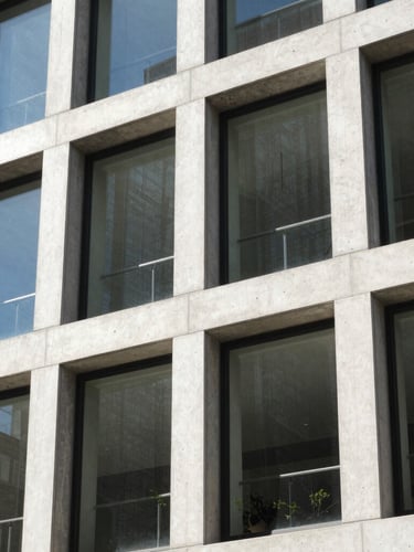 Close-up of a modern building facade in Boise's urban core, showcasing raw concrete, large windows, and sharp geometric shadows. Bright daylight.
