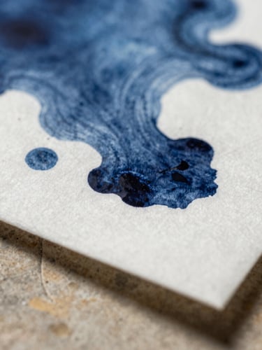 Macro shot of physical indigo ink bleeding into heavy cotton paper, extreme close-up of texture, soft directional side-lighting casting subtle shadows on a warm stone surface.