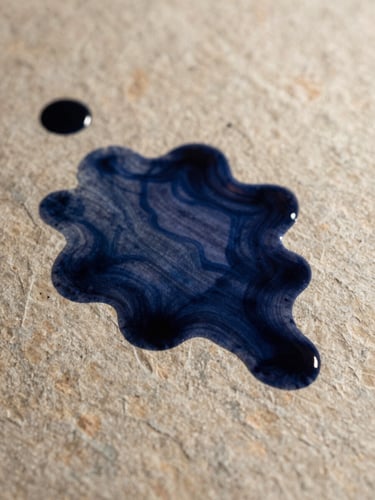 Extreme close-up of dark indigo ink bleeding into heavy, textured warm stone paper, soft directional side-lighting casting subtle shadows.