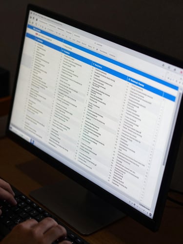 Wide-angle overhead of a risk matrix spreadsheet open on a dark monitor, columns of contract clause categories illuminated by a narrow neon blue highlight bar, cool shadowless studio environment, hands visible at the keyboard edge