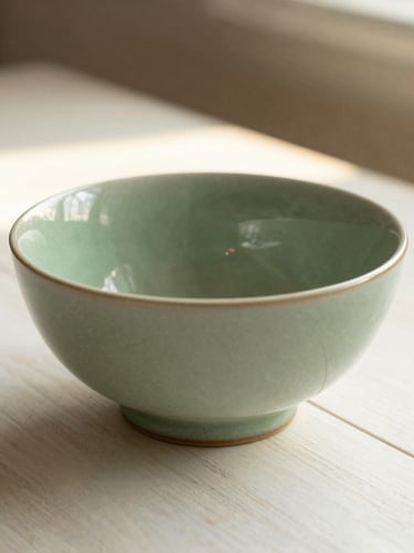 Close-up of a finished sage green ceramic bowl sitting on an almond white wooden table, a single shaft of warm sunlight highlighting the textured glaze, 35mm
