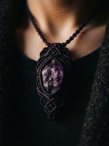 Close-up of a dark macrame talisman with a raw amethyst stone, worn on a person's chest against a dark, textured fabric, moody chiaroscuro lighting, deep purple and black tones, macro lens, high-contrast shadows, shallow depth of field.