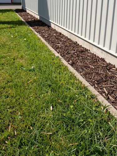 Slightly elevated angle looking down a freshly edged lawn border along a commercial property perimeter, mulch bed running along the building foundation with clean crisp edges, mid-morning natural light casting short shadows, no people, equipment visible in the far background