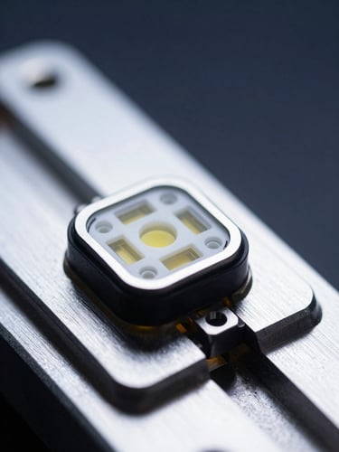 Extreme macro close-up of a compact optical CGM sensor module resting on a precision aluminum fixture, viewed from a slight angle — multiple wavelength emitter apertures visible in sharp detail, shadowless clinical white LED illumination from above, deep navy background fading into the right edge, no hands or people, no text