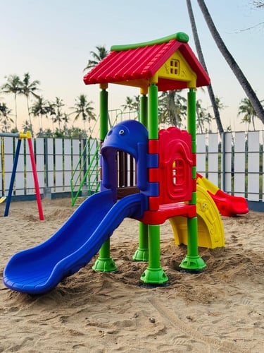 Colorful outdoor plastic playground equipment with a blue slide on a sandy beach under palm trees.