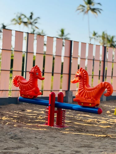 A bright orange seahorse seesaw on a sandy playground with tropical palm trees in the background.