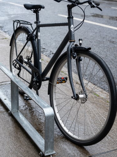 Low-angle street perspective of a utility commuter bike locked to a heavy steel rack on a wet asphalt sidewalk, overcast daylight, water droplets on the frame.