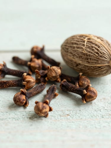 Close-up of dried organic cloves and nutmeg on a rustic wooden surface, soft natural daylight, muted sage green tones, airy editorial photography