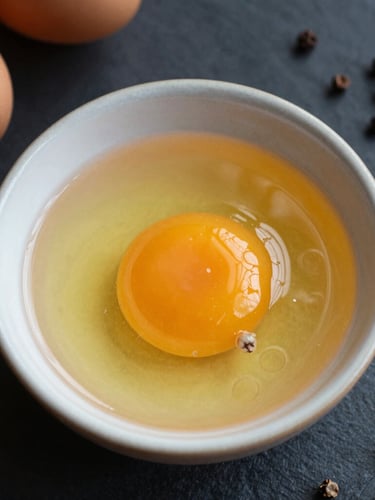 Overhead macro shot of fresh egg yolks in a ceramic bowl next to whole black peppercorns on a dark slate surface, warm morning light, moody editorial photography.