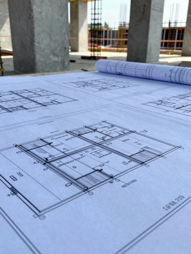 Close-up of civil engineering blueprints laid out on an active construction site, concrete columns and steel reinforcement bars visible in the sharp natural daylight background