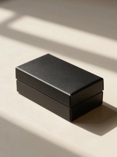 Macro photograph of a minimalist, elegant matte-black box resting on a clean cream ceramic surface, warm natural afternoon sunlight casting long soft shadows, architectural composition.