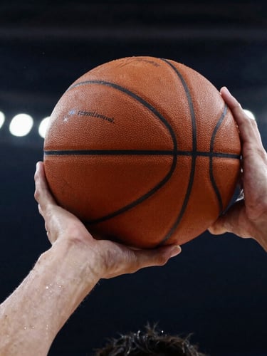 Close-up shot of a male athlete's hands gripping a basketball, sweat glistening under dramatic overhead gym lighting, high-contrast athletic editorial.