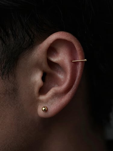 Macro close-up of a delicate gold cartilage piercing on a human ear, elegant minimalist jewelry design, dark moody editorial photography, dramatic chiaroscuro lighting, deep black background, sharp focus, 35mm lens
